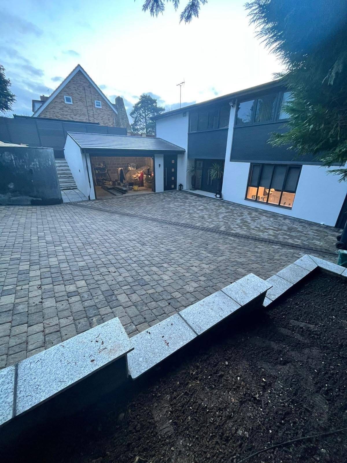 Tumbled sett driveway sweeping up to a rendered house at dusk with the windows lit, edged with light granite kerbs against a newly dug planting bed