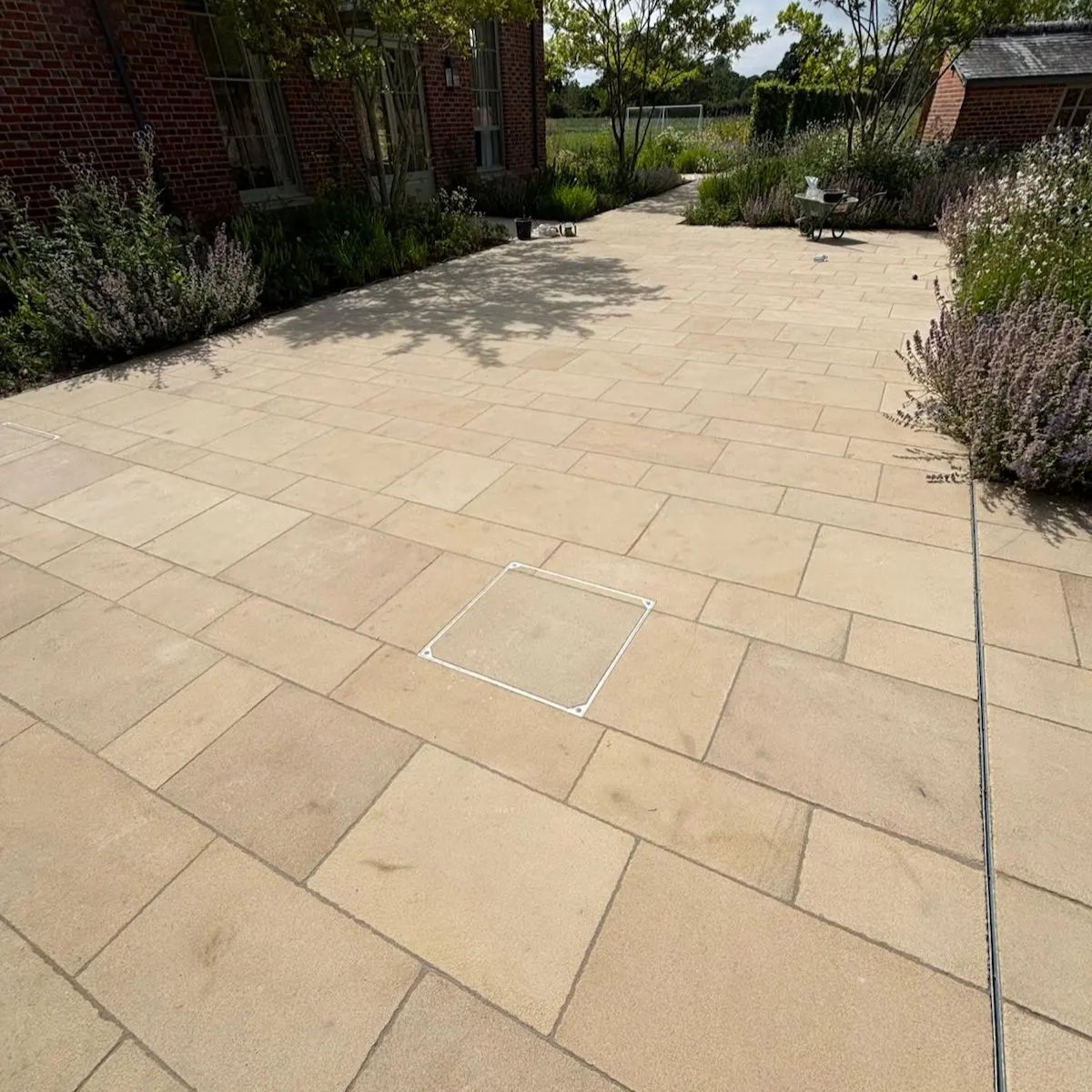 Buff Indian sandstone patio laid in a random rectangular pattern beside a red-brick house, with a recessed manhole cover and lavender planting